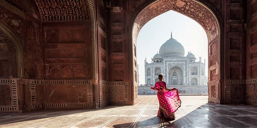 A woman in a beautiful pink sari looks through an ornate archway to see the iconic Taj Mahal. 