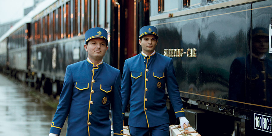 Two members of Venice Simplon-Orient-Express staff, dressed in their iconic blue uniform, carry luggage on the train platform. 